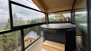 Uma banheira de hidromassagem moderna numa varanda, com amplas janelas que oferecem vista para uma área montanhosa coberta de árvores.