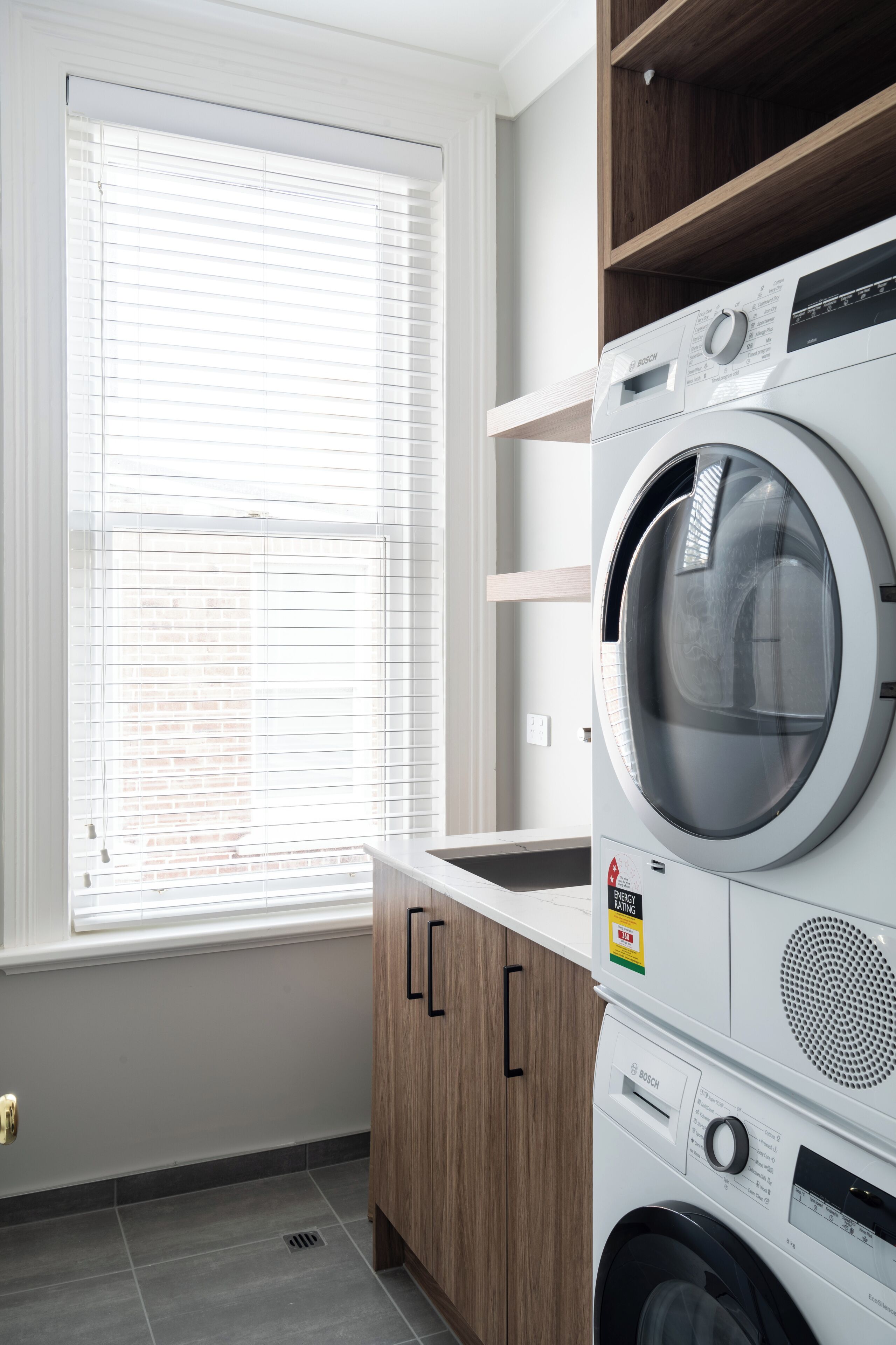A compact laundry room with a Bosch washing machine, wooden cabinets, and a window with blinds.