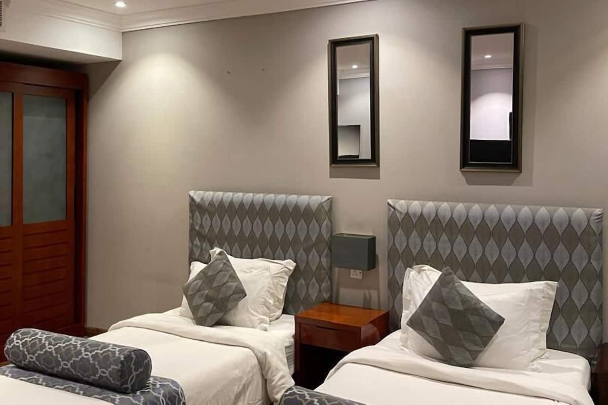 Ein Hotelzimmer mit zwei Betten, jedes mit gemusterten Kissen und Kopfteilen, einem Nachttisch mit Lampe und einem Spiegel an der Wand.