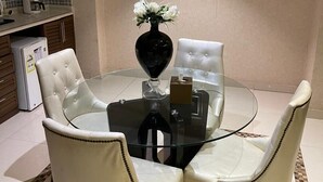 Ein Essbereich mit einem Glastisch, um den vier weiße Ledersessel stehen. Auf dem Tisch steht eine Vase mit weißen Blumen, im Hintergrund ist ein kleiner Kühlschrank zu sehen.