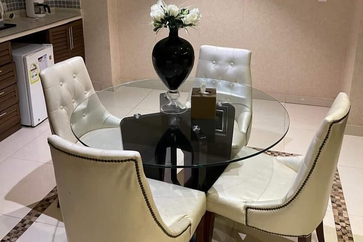 Ein Essbereich mit einem Glastisch, um den vier weiße Ledersessel stehen. Auf dem Tisch steht eine Vase mit weißen Blumen, im Hintergrund ist ein kleiner Kühlschrank zu sehen.