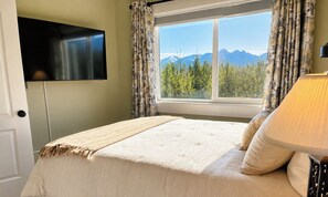Ein Schlafzimmer mit einem Bett, einem an der Wand montierten Fernseher, einem Fenster mit Blick auf Berge und Vorhängen.