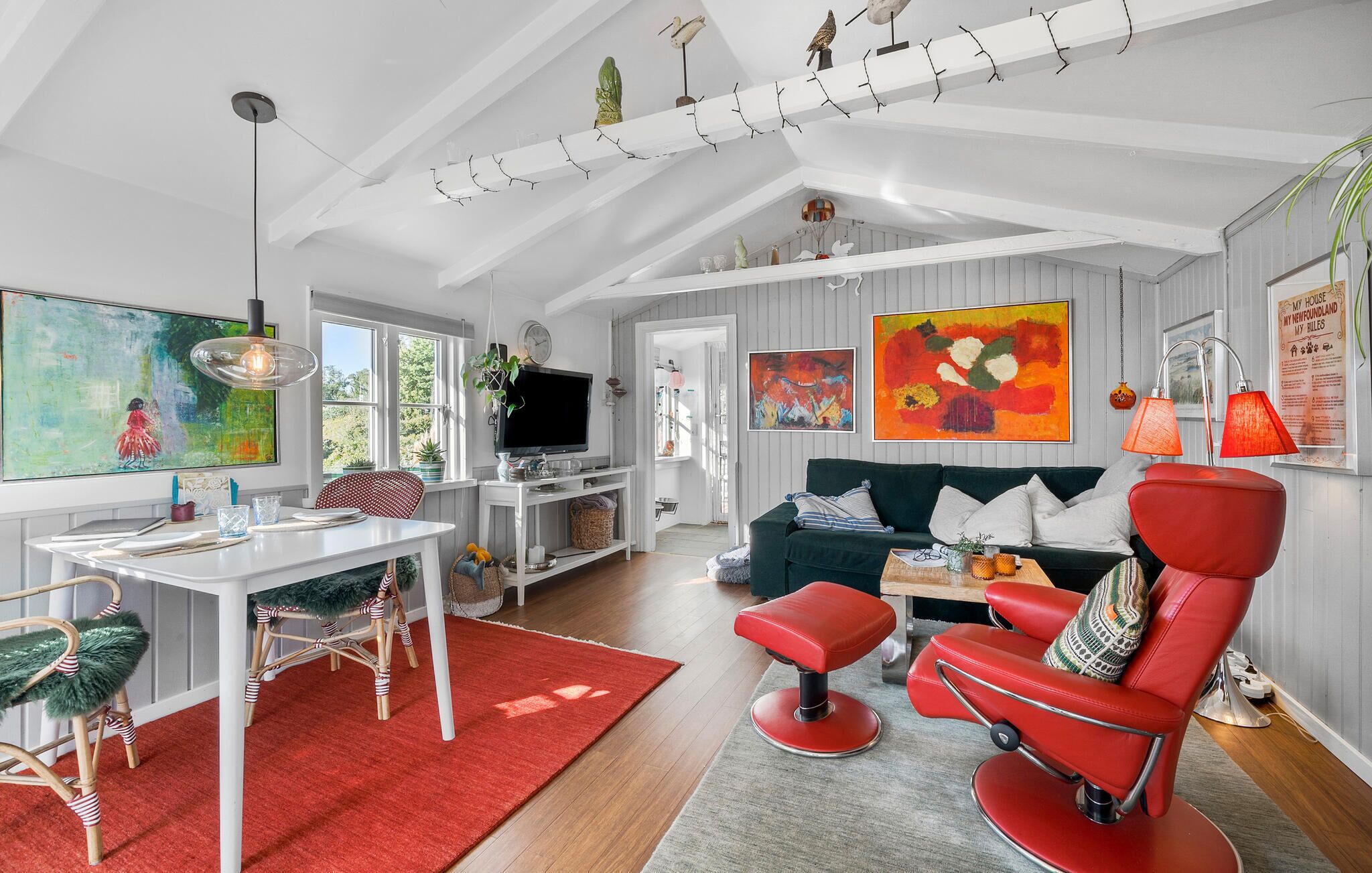 A modern living room with a TV, sofa, and red chairs. There are paintings on the walls and a window with a view of trees.