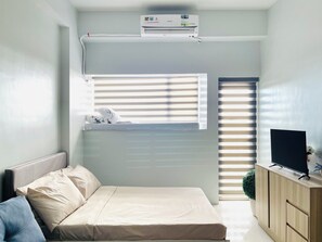 A compact bedroom with a bed, a mounted air conditioner, a window with blinds, a TV on a wooden cabinet, and a small plant. - Hubomoto WV Suites (Iloilo)