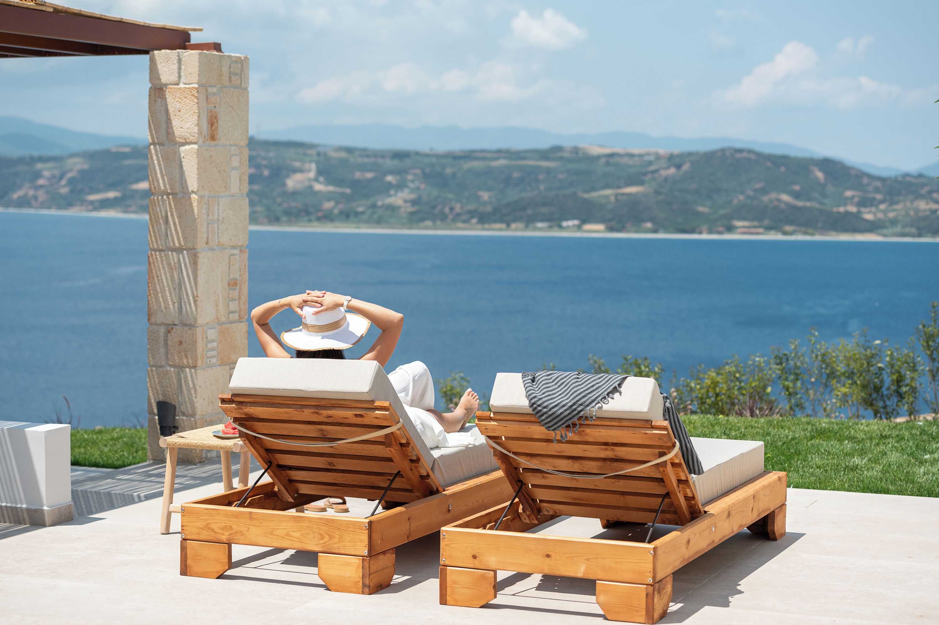 Two wooden lounge chairs with cushions, a stone pillar, and a view of the sea and hills.