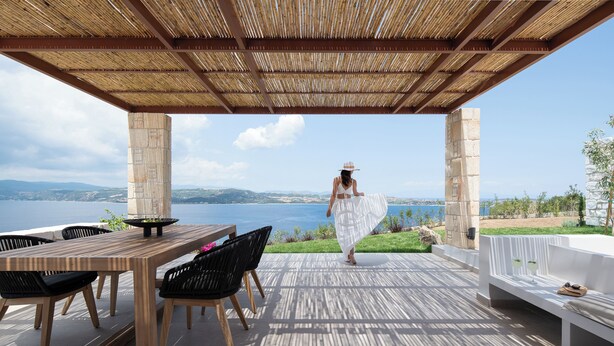 Patio esterno con tavolo e sedie di legno, una panca bianca e vista sul mare.