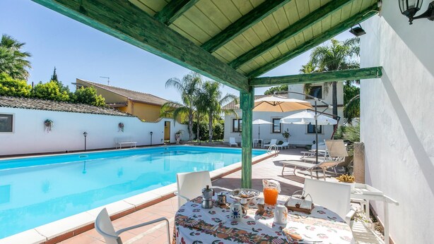 A poolside area with a table set for breakfast, a covered seating area, and a view of a white building with a tiled roof.