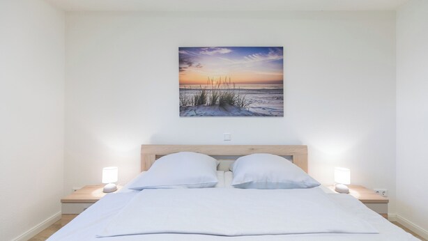 A bedroom with a bed, two bedside tables, and a wall-mounted picture of a beach scene. - Ferienwohnung Schöne Aussicht in Emmelsbüll-horsbüll (Emmelsbüll-Horsbüll)