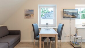 A modern living room with a sofa, a dining table with chairs, a TV mounted on the wall, and a window with a view of the outdoors.