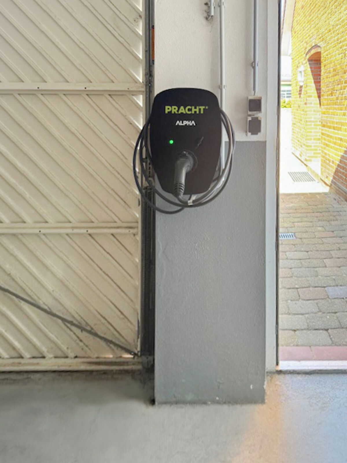 A charging station labeled "PRACT ALPHA" mounted on a wall near a garage door.