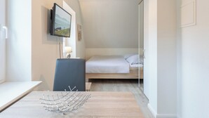 A small, modern room with a bed, a desk, and a chair. There is a TV mounted on the wall and a lamp on the desk.