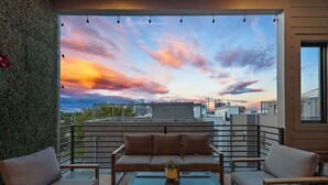 Una terraza en la azotea con sofá, sillas, una mesa pequeña y vista del horizonte de la ciudad al atardecer.