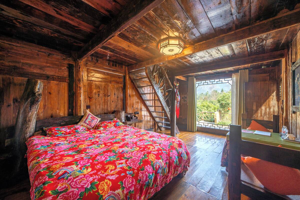 Interior de una cabaña de madera con cama, escalera y vistas por una ventana.