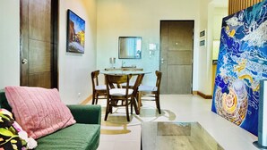 A living room with a green sofa, a coffee table, and a dining area with a table and chairs. There is a painting on the wall and a door leading to another room. - Hubomoto Megaworld Suites (Iloilo)
