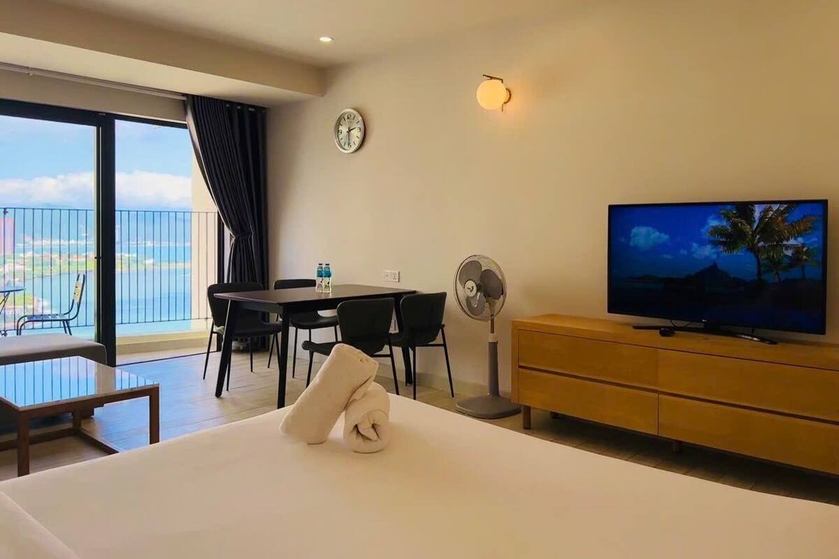 A hotel room with a bed, a TV, a dining table with chairs, a fan, a clock, and a balcony with a view of the sea.