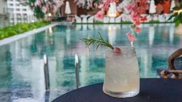 A glass of a clear beverage with a rosemary sprig on a table by a pool with a floral backdrop.