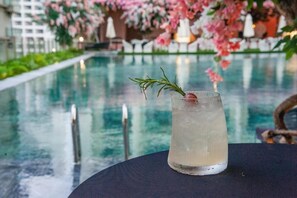 Ein Glas mit einem klaren Getränk und einem Rosmarinzweig auf einem Tisch neben einem Pool, mit floralem Hintergrund.