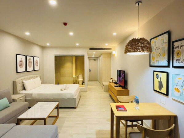 A modern hotel room with a bed, sofa, and a dining area. There are framed pictures on the wall, a TV, and a water bottle on the table.