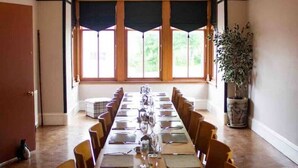 Une salle de conférence avec une longue table préparée pour une réunion, de grandes fenêtres avec des rideaux, un lustre et une plante en pot.