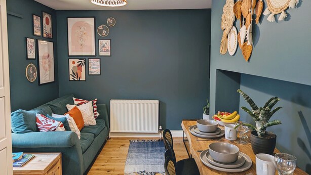 Un salon chaleureux avec un canapé vert, une table à manger dressée avec des assiettes et des tasses, et diverses œuvres d’art accrochées aux murs.