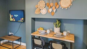 Un coin repas avec une table en bois dressée pour deux personnes, un téléviseur fixé au mur et une décoration murale faite de feuilles tressées.