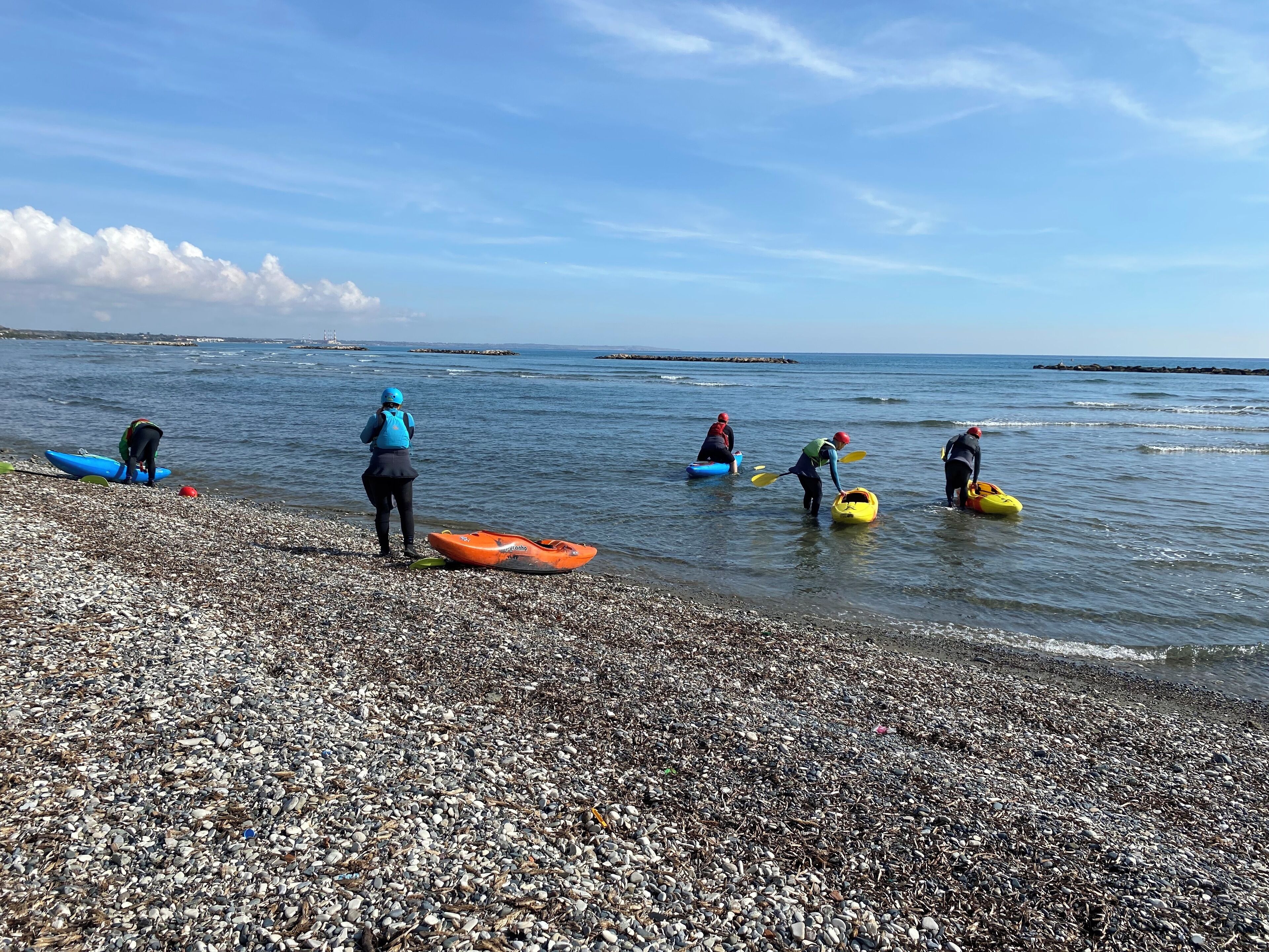 A pebbled beach with kayaks and people preparing for water activities.
