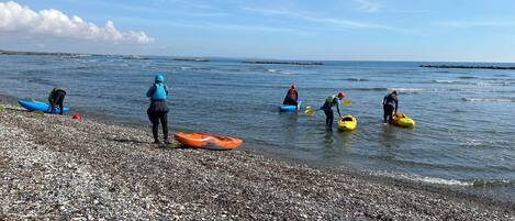 A pebbled beach with kayaks and people preparing for water activities.