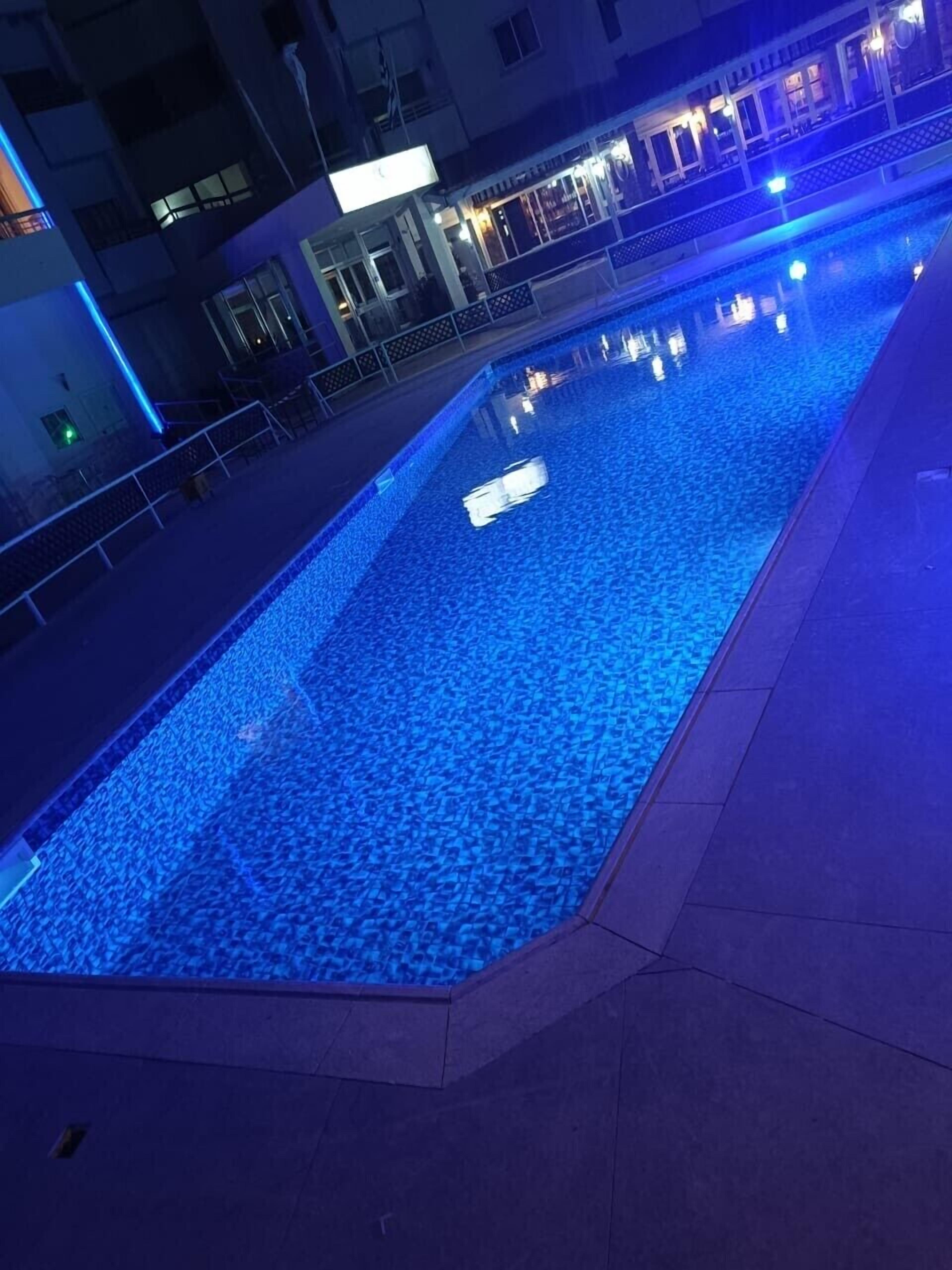 A swimming pool with blue lighting, surrounded by a building with balconies and a railing.