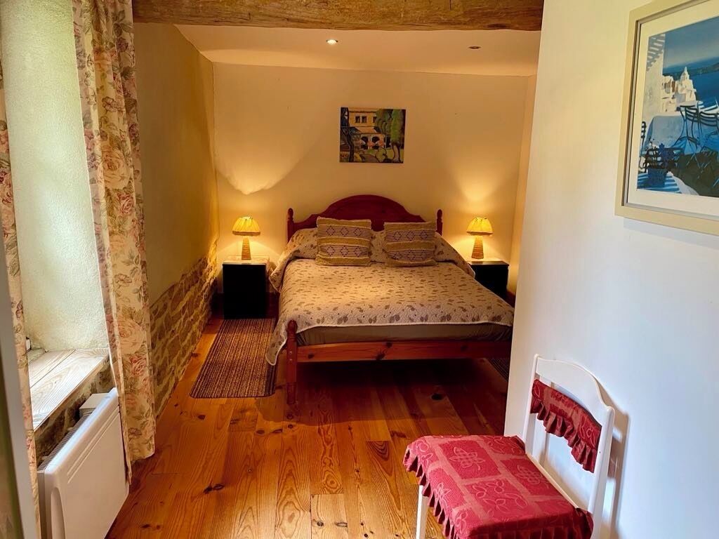 A cozy bedroom with a wooden floor, a bed with patterned bedding, two bedside lamps, and a framed picture on the wall.