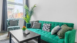 Uma sala de estar com um sofá verde, uma mesinha com um controle remoto e um vaso de planta.