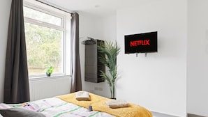 Quarto com cama, janela com cortinas, planta em vaso, televisão na parede e um cabideiro.