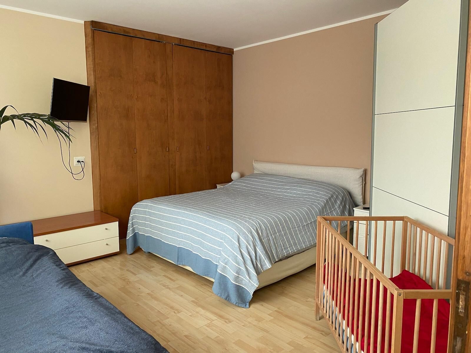 A bedroom with a bed, a wooden wardrobe, a desk, a chair, and a crib.