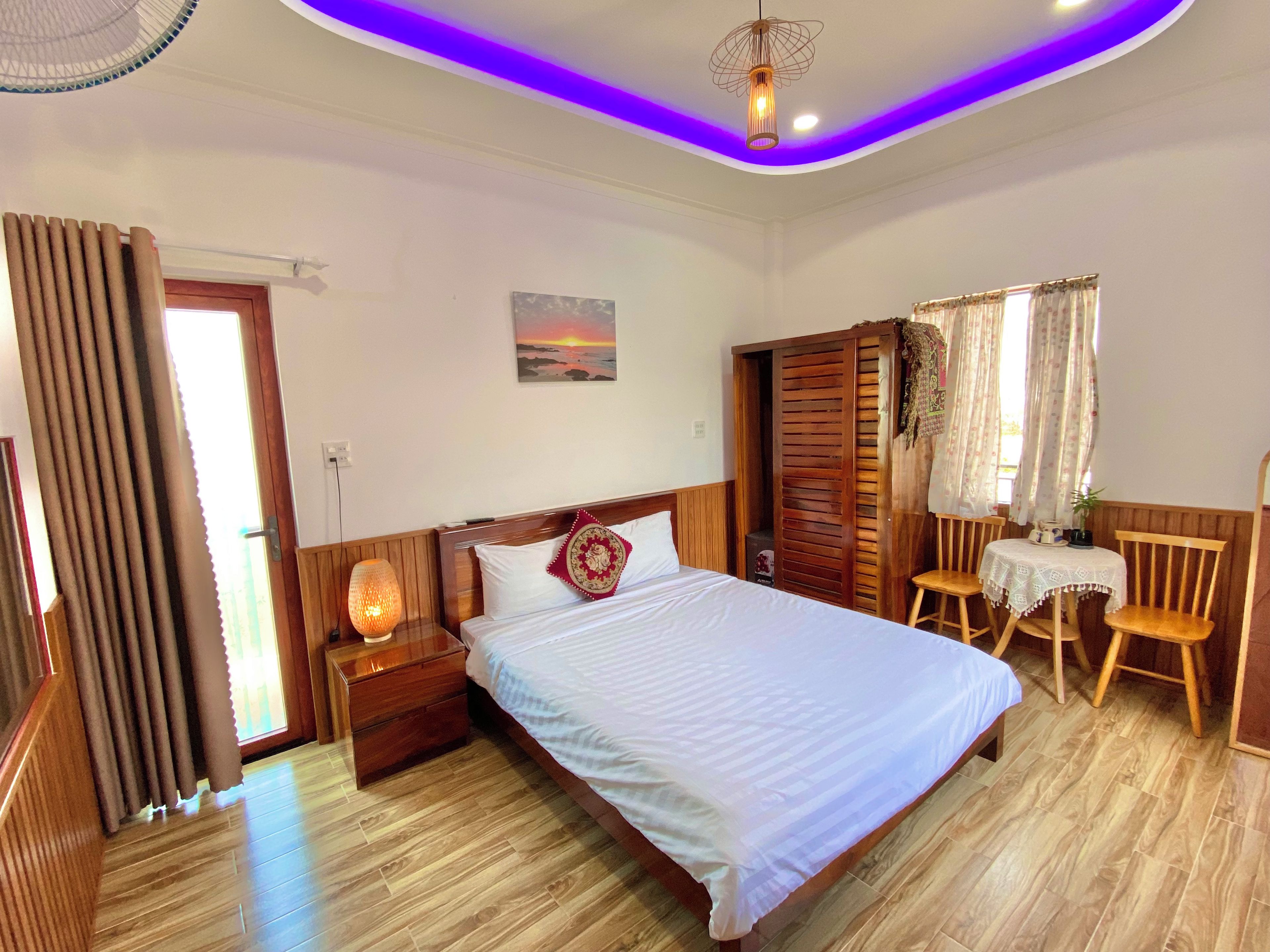 Una habitación de hotel con una cama, cabecera de madera, mesita de noche con una lámpara, una pequeña mesa con sillas y una ventana con cortinas.
