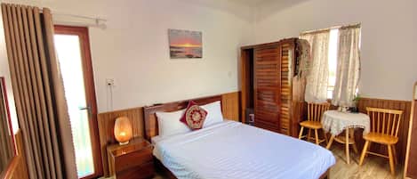 Une chambre d’hôtel avec un lit, une tête de lit en bois, une table de chevet avec une lampe, une petite table avec des chaises et une fenêtre avec des rideaux.