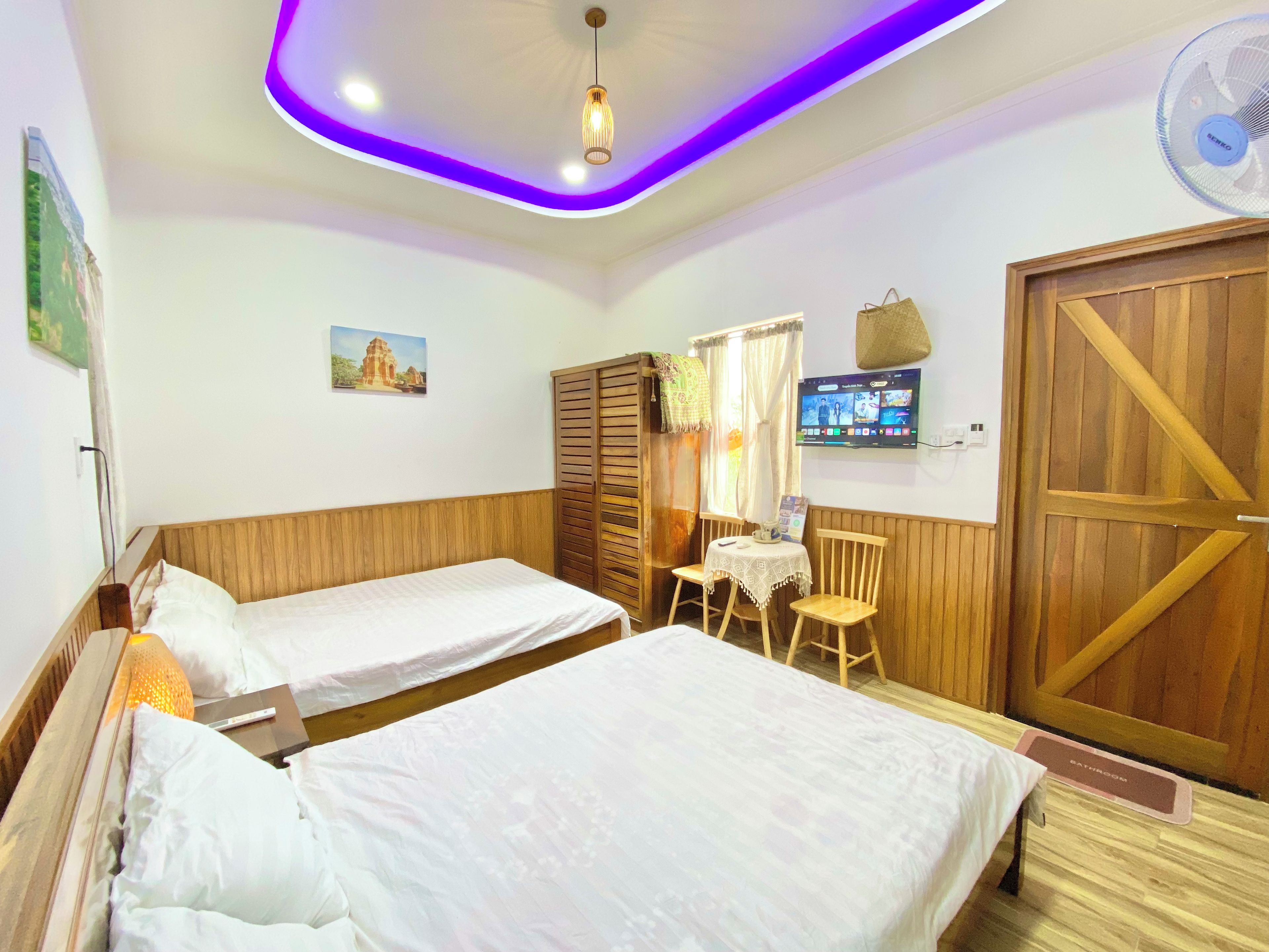 A room with two beds, a wooden wardrobe, a television on the wall, and a ceiling with purple lighting.