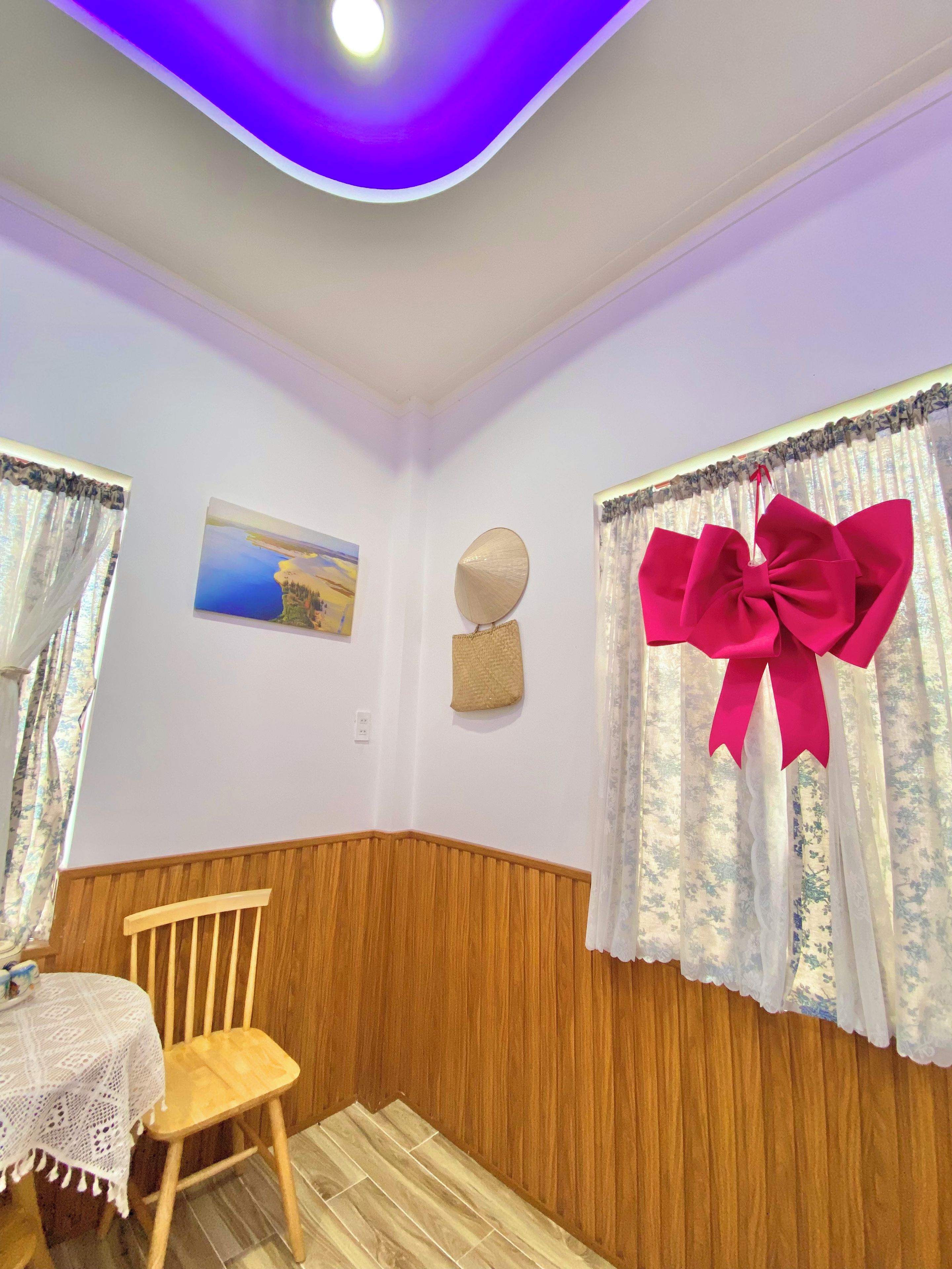 Une pièce avec une chaise en bois, une table recouverte d’une nappe en dentelle, un tableau, un élément décoratif mural et un grand nœud sur le rideau de la fenêtre.