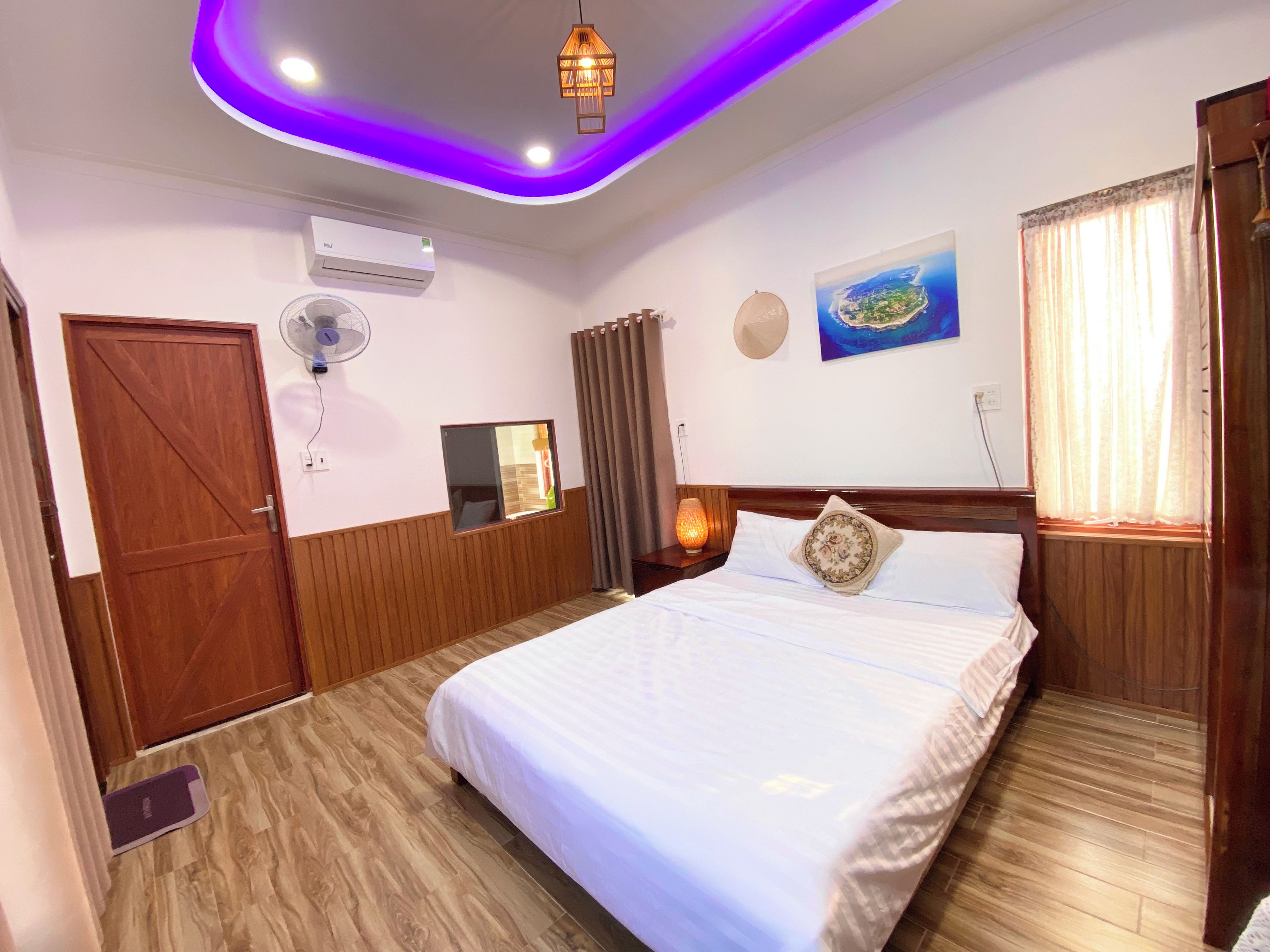 A hotel room with a bed, wooden paneling, a ceiling fan, an air conditioning unit, a window with curtains, and a wall-mounted lamp.