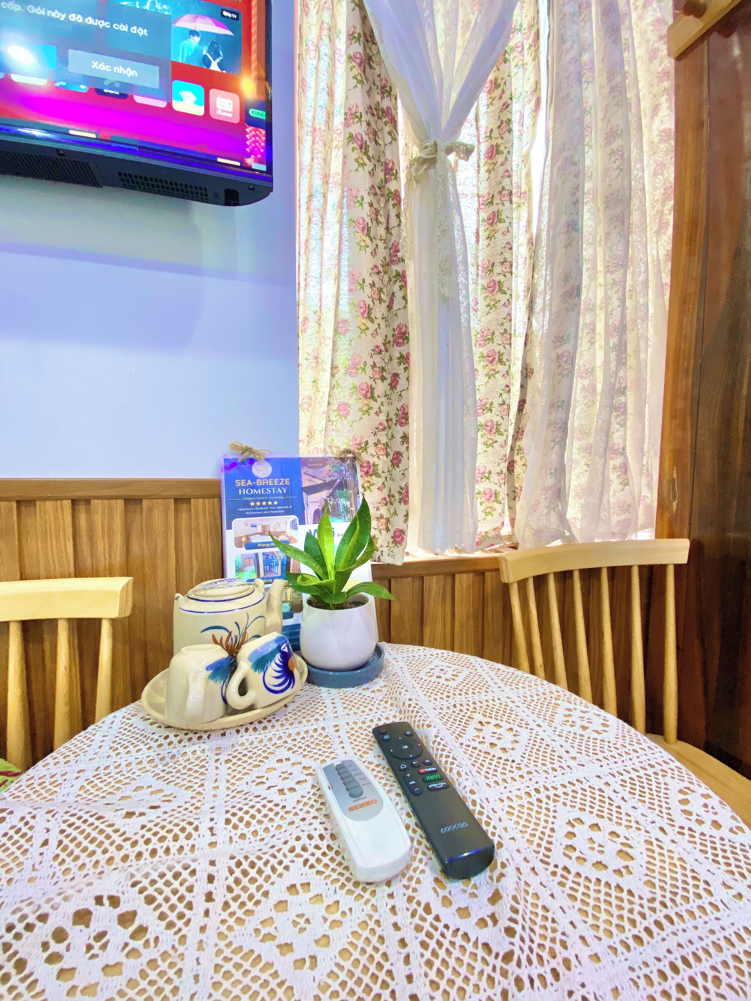 Una habitación con una televisión montada en la pared, una mesa con mantel de encaje, un control remoto, una tetera, una taza y una planta en maceta.