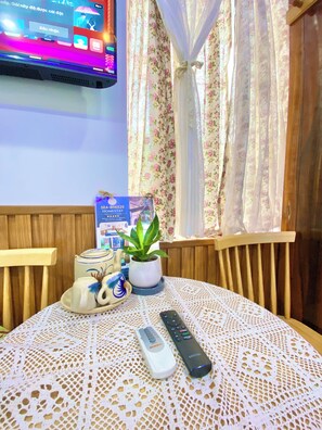 A room with a television mounted on the wall, a table with a lace tablecloth, a remote control, a teapot, a cup, and a potted plant. - Sea-breeze homestay (Phu Thuy)