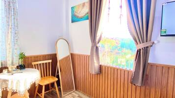 A room with a bed, a wooden chair, a small table with a plant, and a window with curtains.