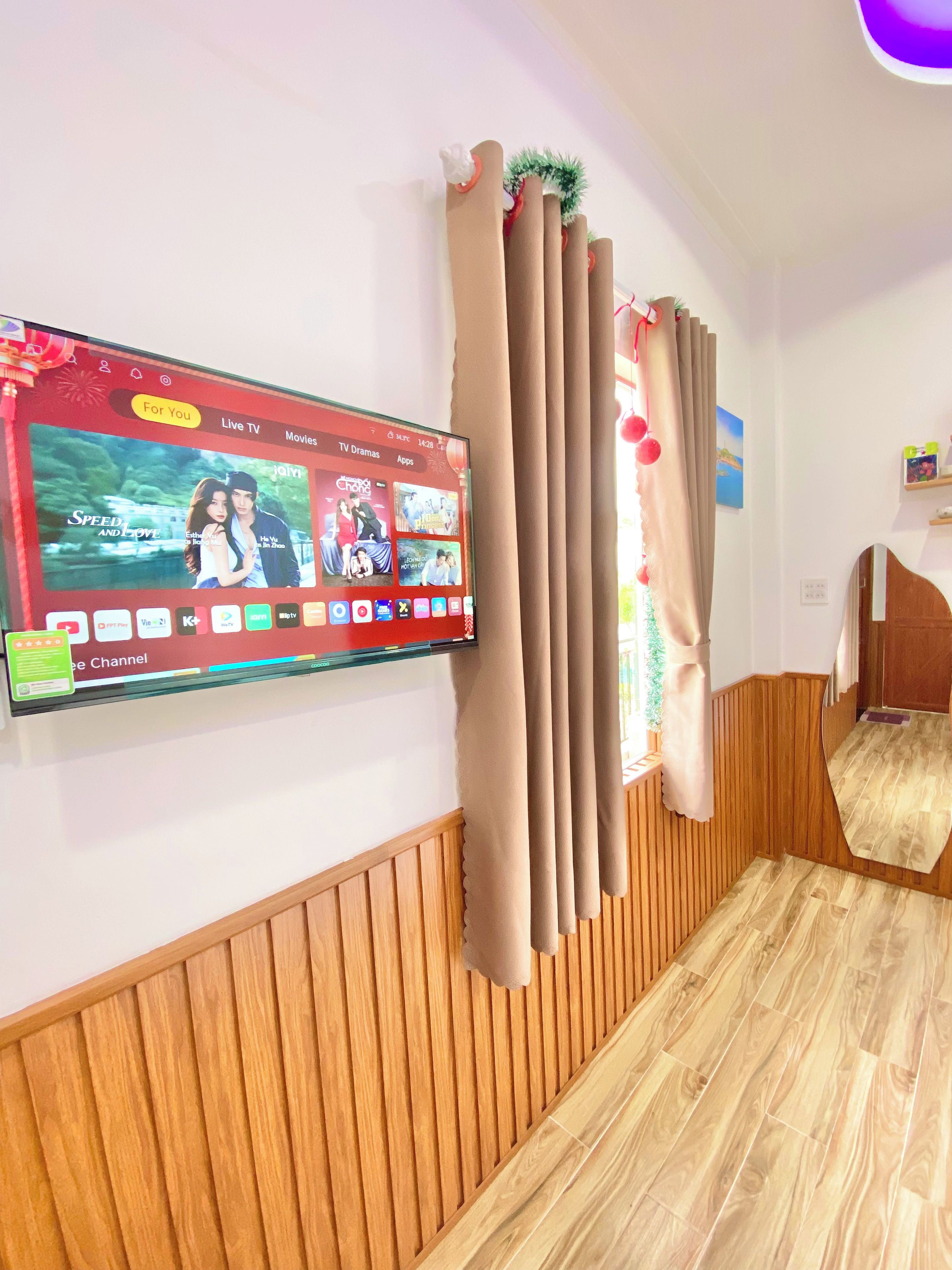 A room with a mounted TV displaying a live TV channel interface, wooden paneling on the walls, and a window with curtains.