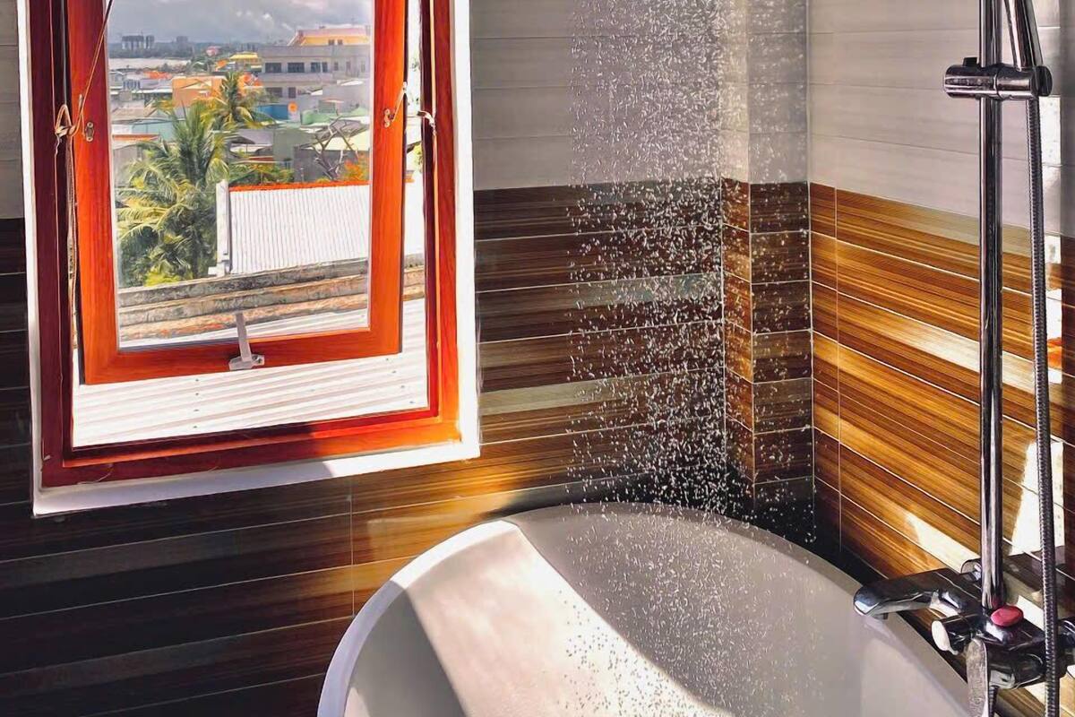 Un bagno con una vasca, un soffione doccia e una finestra vista edifici e alberi.