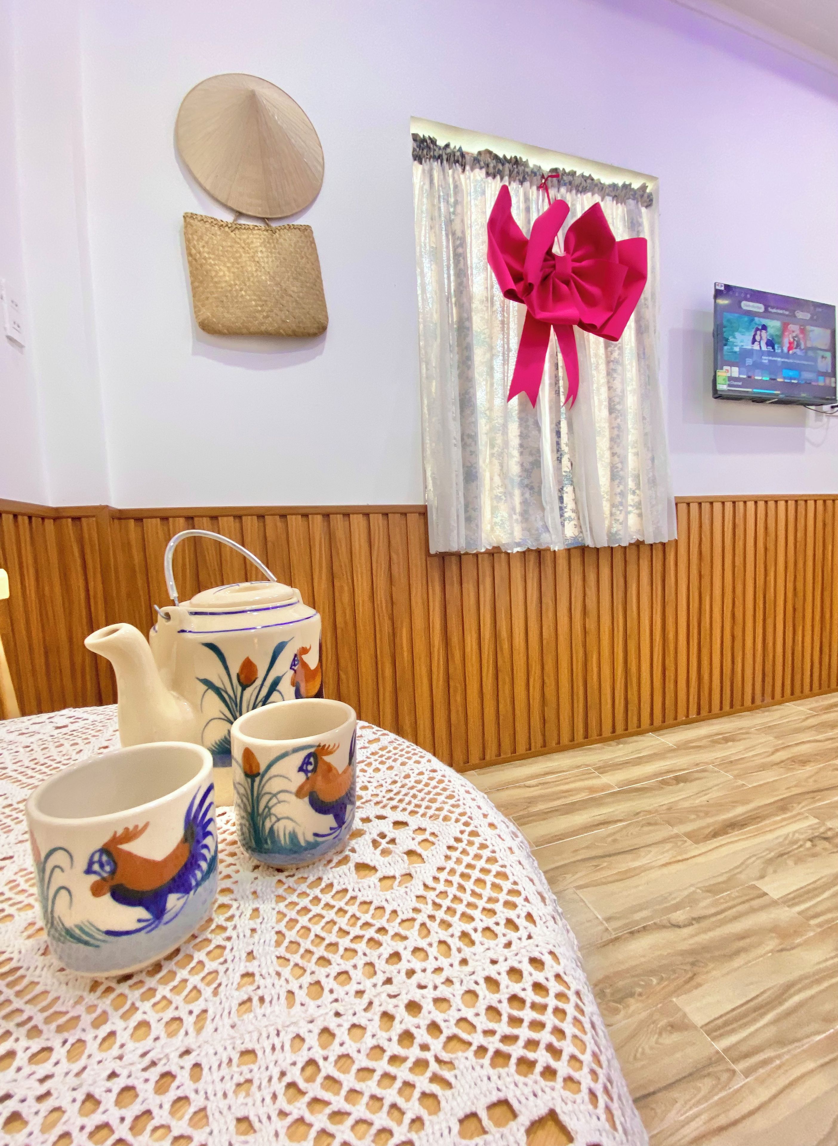 A room with a wooden paneled wall, a table with a crocheted tablecloth, a teapot, two cups, a decorative hat, and a curtain with a large bow.
