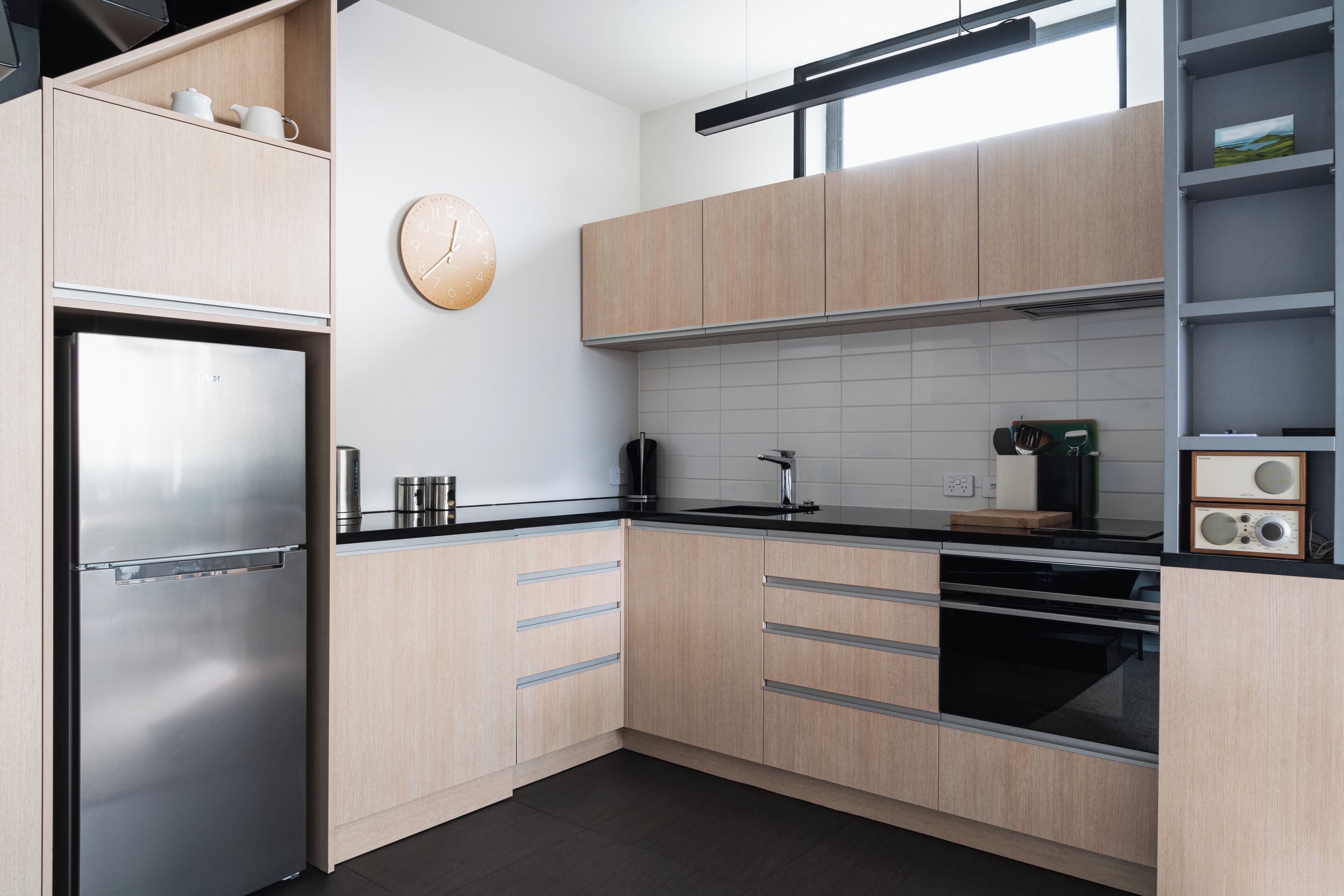 Una cucina moderna con pensili in legno chiaro, un frigorifero in acciaio inox, un piano di lavoro nero, una sveglia al muro e una radio sullo scaffale.