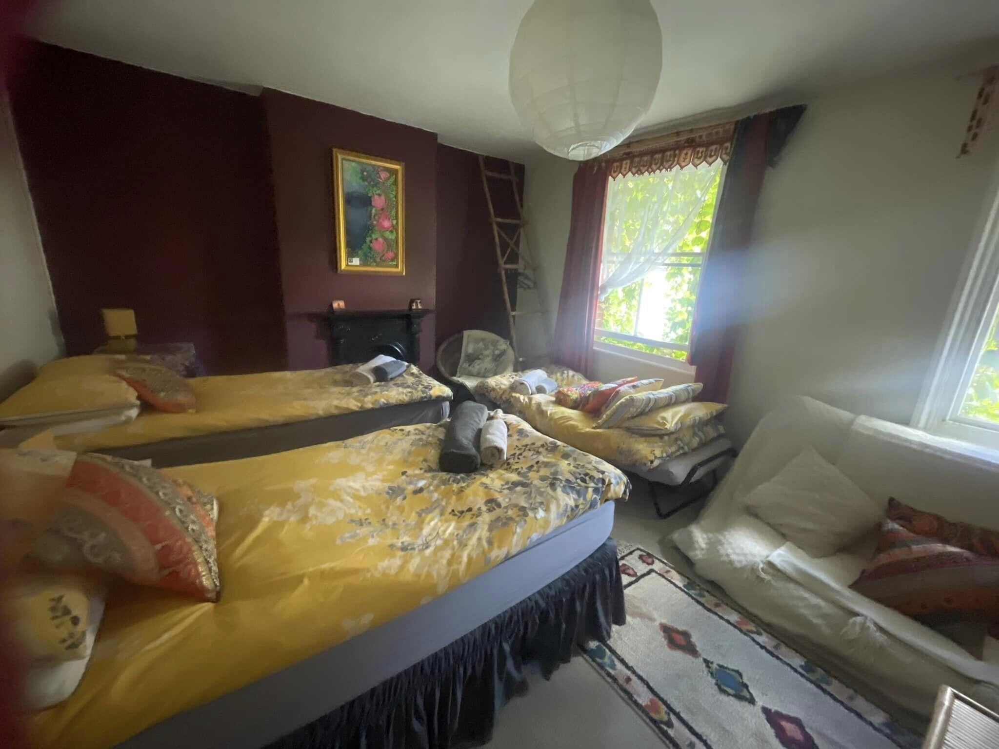 A room with two beds, a chair, a ladder, a painting, and a window with curtains.