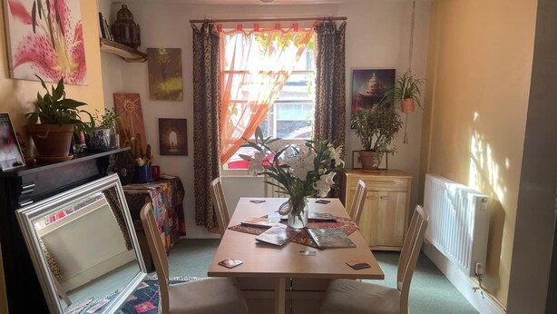 Uma área de refeições com uma mesa posta para dois, cercada por cadeiras. Há plantas em vasos no parapeito da janela e um radiador na parede.