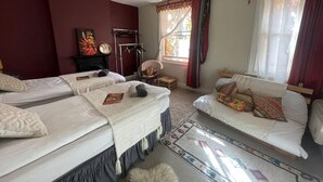 Quarto com duas camas, um sofá, uma mesinha e uma cadeira. Há uma janela com cortinas e um lustre.