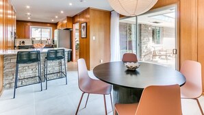 A modern kitchen with wooden cabinets, a central island with bar stools, and a dining area with a round table and chairs. - Desert Rose: 1950s southwest modern home (Tulsa)