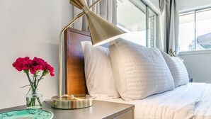 A neatly made bed with white bedding, a bedside lamp, a vase with flowers, and a plate on a bedside table. - Desert Rose: 1950s southwest modern home (Tulsa)