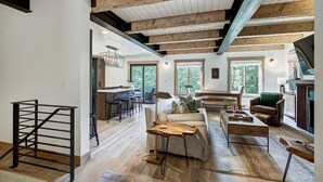 A spacious living area with a high ceiling, exposed wooden beams, a central seating area with a sofa and coffee table, and a kitchen area with a bar and stools.